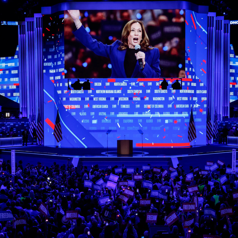 fotografija iz 2024 DNC gomile koja gleda monitor na kojem se vidi Kamala Harris kako govori u mikrofon s jednom podignutom rukom...
